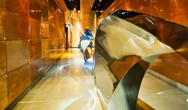 Corredor interno revestido de superfícies metálicas douradas e brilhantes, com iluminação quente. No centro, há uma grande escultura abstrata de metal polido, de formas curvas e orgânicas, que reflete o ambiente ao redor.