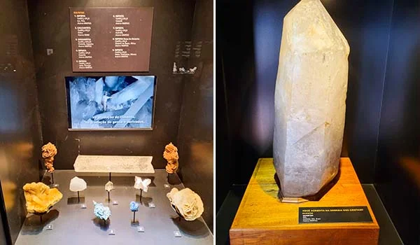 Fotografia de uma exposição de minerais e pedras em um museu. À esquerda, uma vitrine iluminada apresenta diversos minerais e cristais de diferentes formas e cores, acompanhados de placas explicativas e uma tela com vídeo. À direita, destaca-se um grande cristal ou mineral branco, translúcido e de formato alongado, exposto verticalmente sobre uma base de madeira.