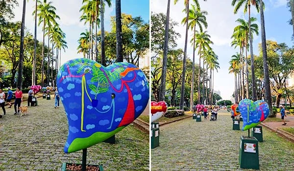 Fotografia de uma praça arborizada, com uma fileira de palmeiras altas e calçamento de pedras. Em primeiro plano, há uma grande escultura colorida de um coração, pintada com elementos de natureza e apoiada sobre uma base. Ao longo da praça, outras esculturas semelhantes aparecem em sequência, enquanto pessoas caminham e observam as obras.