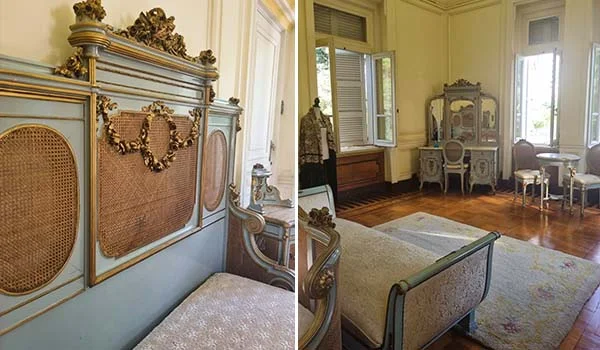 Fotografia de um quarto de estilo clássico e luxuoso, com móveis antigos em tons de azul-claro e detalhes dourados. À esquerda, destaca-se a cabeceira ornamentada de uma cama, com painéis de palhinha e entalhes decorativos. Ao centro e à direita, há uma penteadeira com espelho, cadeiras e outros móveis de época. O ambiente possui piso de madeira polida, paredes claras com molduras decorativas, janelas altas com persianas e um grande tapete claro. Visita ao Palácio da Liberdade.