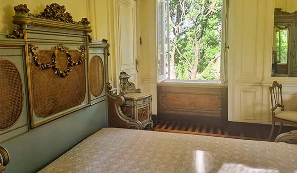 A imagem mostra um quarto do Palácio da Liberdade, em Belo Horizonte (MG), com mobiliário de estilo histórico, incluindo uma cama de madeira ricamente ornamentada, mesa de cabeceira e grandes janelas voltadas para a área externa.
