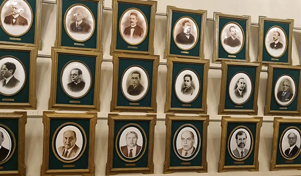 Parede interna com uma coleção de retratos antigos em preto e branco de homens, dispostos em molduras douradas e verdes, organizadas em três fileiras. Os retratos parecem representar antigos governadores ou autoridades.