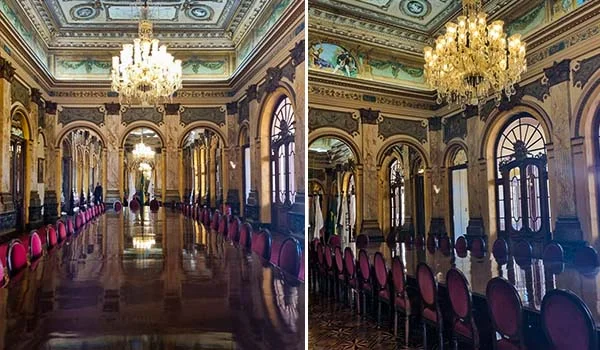Salão luxuoso e ornamentado, com longas mesas de madeira polida cercadas por cadeiras estofadas em vermelho. O ambiente possui grandes espelhos e portas e janelas arqueadas, paredes decoradas com detalhes dourados e pinturas, além de um teto ricamente trabalhado. Grandes lustres de cristal iluminam o salão, cujo piso brilhante reflete a decoração. Visita ao Palácio da Liberdade.