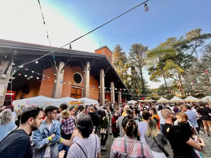 magem de uma festividade ao ar livre com muitas pessoas reunidas em frente a um espaço cultural, decorado com luzes e bandeirinhas coloridas, em um dia ensolarado com árvores ao fundo.