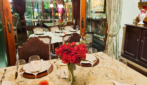 Foto de uma bonita mesa posta com arranjo de flores vermelhas ao centro, no no entorno móveis e outra mesa posta. Uma das dicas sobre onde comer na Serra da Mantiqueira.