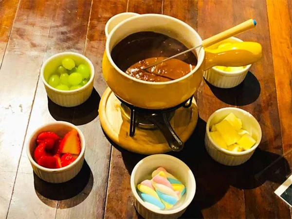 Foto com uma vasilha típica de fondue de chocolate, tendo ao redor diversos podes com frutas e marshmallow, dicas sobre onde comer na Serra da Mantiqueira.