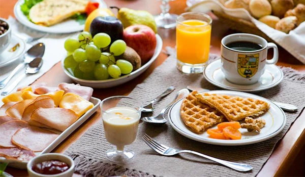Uma mesa farta com um delicioso café da manhã contendo waffles, frutas, presunto, chá e suco de laranja em um ambiente acolhedor.