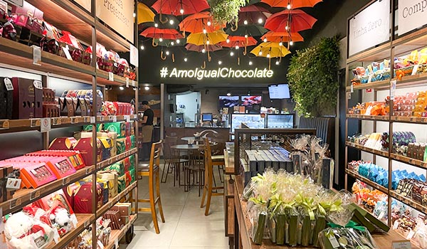 Interior de uma loja de chocolates, com prateleiras recheadas de produtos e decoração alegre com sombrinhas coloridas. O ambiente convida a apreciar o chocolate.