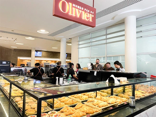 Imagem da padaria Mundo Pão do Olivier, mostrando uma variedade de doces e salgados em uma vitrine com clientes e funcionários ao fundo.