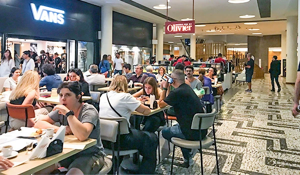 Interior do Conjunto Nacional com várias pessoas em mesas, lojas ao fundo mostrando um ambiente movimentado e moderno.