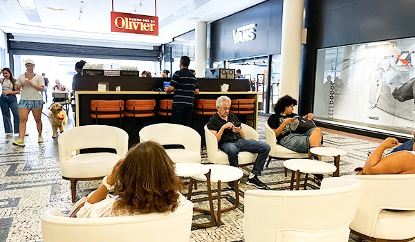 Imagem de um Café Olivier, onde pessoas estão sentadas conversando e utilizando dispositivos móveis. Existem mesas, cadeiras e um cachorro ao fundo.