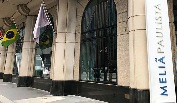 Fachada do hotel Meliá Paulista, com grandes portas de vidro, colunas de pedra, bandeiras do Brasil e uma placa vertical com o nome “Meliá Paulista”.
