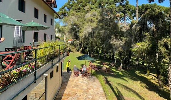 Foto da área externa do hotel com árvores, céu azul, mesa com cadeiras com guarda-sol e turistas sentados.