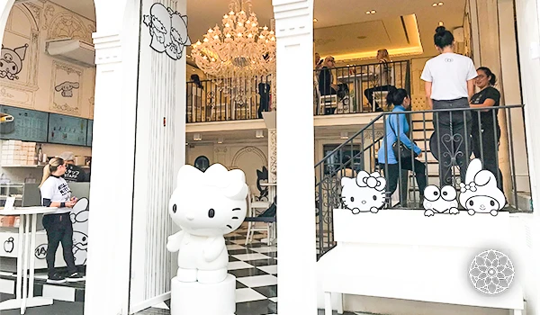 Interior de cafeteria decorada com temática Hello Kitty, destaque para figura gigante da Hello Kitty ao lado de crianças e adultos interagindo, decoração com personagens fofos e ambiente acolhedor