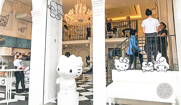 Imagem de um espaço de entretenimento com decoração temática de Hello Kitty, incluindo uma estátua da personagem, pessoas jovens e ambiente decorado com detalhes fofos.