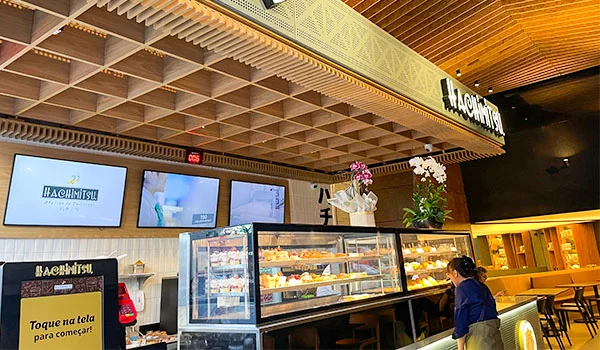 Interior da Cafeteria Hachimitsu, com balcão refrigerado repleto de doces e sobremesas, plantas decorativas e telas exibindo imagens de produtos. Ao fundo, há um painel com o nome da confeitaria e mesas para atendimento.