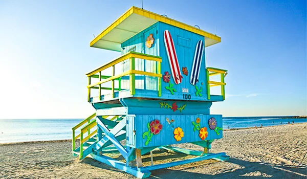 Posto de salva-vidas colorido em uma praia, construído sobre uma estrutura elevada. A cabine é azul com detalhes amarelos e está decorada com flores e pranchas de surfe pintadas nas paredes. Uma escada lateral leva ao piso superior, cercado por corrimãos. Ao fundo, há uma extensa faixa de areia, o mar e um céu azul sem nuvens.