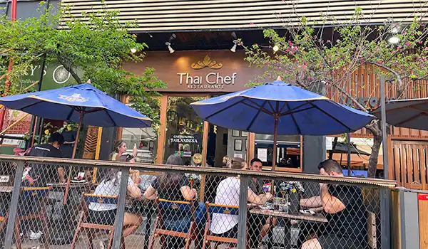 Clientes desfrutando de uma refeição ao ar livre na varanda do restaurante Thai Chef, com árvores e guarda-sóis azuis, criando um ambiente acolhedor e social.