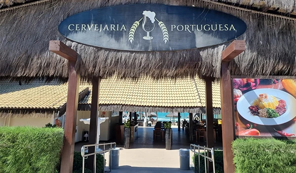 Entrada de uma cervejaria portuguesa com sinal, que exibe o nome 'Cervejaria Portuguesa'. A estrutura de madeira com teto de palha oferece um ambiente rústico e acolhedor.