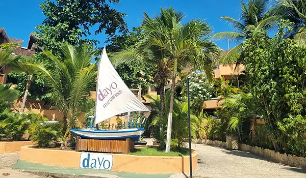 Cenário tropical com árvores palmadas, um barco decorativo e uma bandeira com o logo 'dayo' em um ambiente ao ar livre.