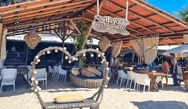 Imagem de um restaurante à beira-mar com espaço ao ar livre, cadeiras de balanço de corda, decoração rústica e um letreiro de madeira indicando 'Cavo Restaurante Hotel'. Atmosfera ensolarada e relaxante na praia.
