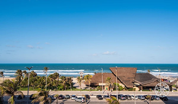 Praia com areia, mar azul e céu claro, com palmeiras e edifícios ao fundo. Perfeita para relaxar e aproveitar o clima tropical.