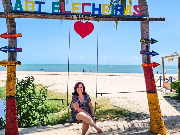 Praia com sinalização de Abert Lecheras e uma mulher sentada em um balanço na areia, céu azul, mar calmo e ambiente tropical.