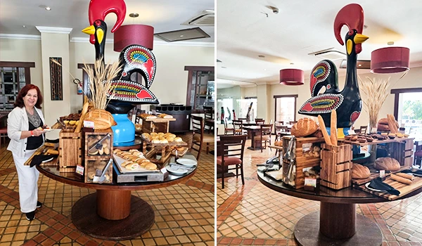 Estampa de buffet de café da manhã com expositor de escultura de galo decorativo em restaurante. Ambiente aconchegante com mesas e iluminação moderna.