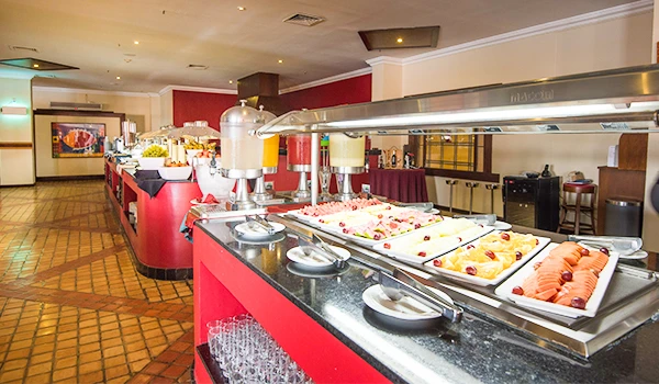 Buffet de café da manhã com diversas opções de alimentos em um restaurante bem decorado, incluindo frutas, pães, queijos e alimentos quentes. Experiência de hospedagem no Vila Galé Fortaleza.
