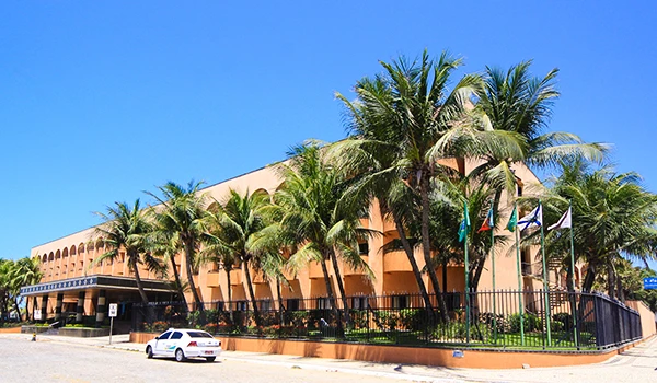 Imagem de um prédio de arquitetura moderna cercado por palmeiras, sob um céu azul claro, em uma área urbana bem cuidada, com uma rua movimentada ao redor. Experiência de hospedagem no Vila Galé Fortaleza.