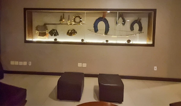 Imagem de uma sala de estar moderna com uma parede de vidro que exibe coleções decorativas, incluindo coroas, objetos de arte e uma guirlanda, além de dois pufes pretos na frente.