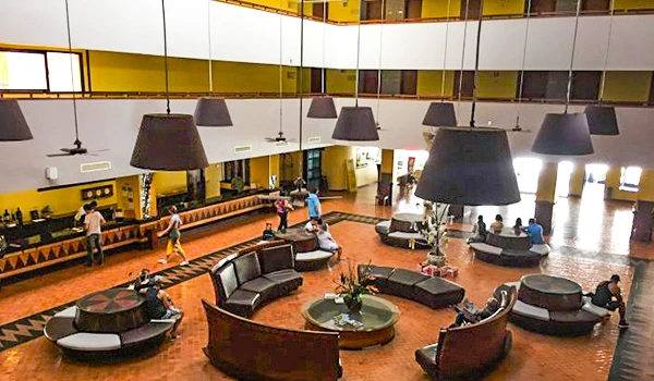 Imagem de um lobby de hotel com mobília moderna em tons escuros e arena de recepção, com várias pessoas relaxando e interagindo. Experiências de hospedagem no Vila Galé Fortaleza.