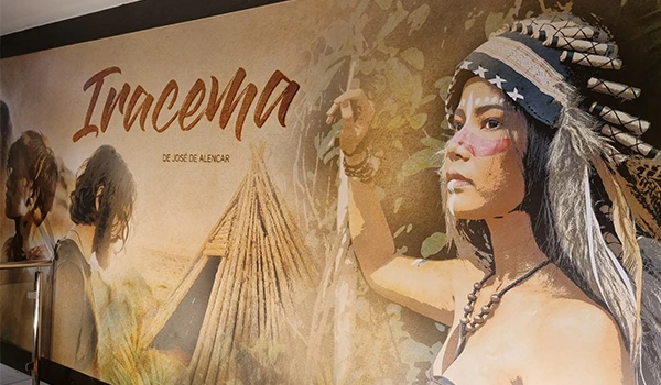 Cenário com tela artística sobre 'Iracema' de José de Alencar, com arte indígena, calçados rústicos e imagens de uma mulher indígena como tema principal. Experiência de hospedagem no Vila Galé Fortaleza.
