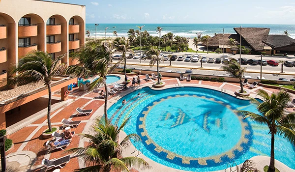 Vista do resort tropical com piscina e vista para o mar, cercado por coqueiros, com área de lazer, estacionamento e uma cabana ao fundo.