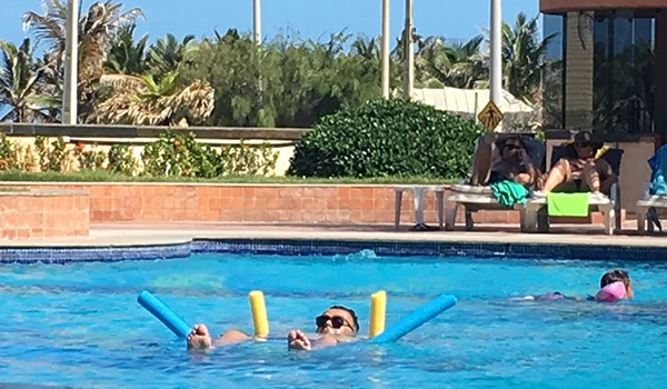 Pessoa relaxando na piscina em um resort de praia, usando óculos de sol e flutuadores de piscina, com ambiente tropical ao fundo.
