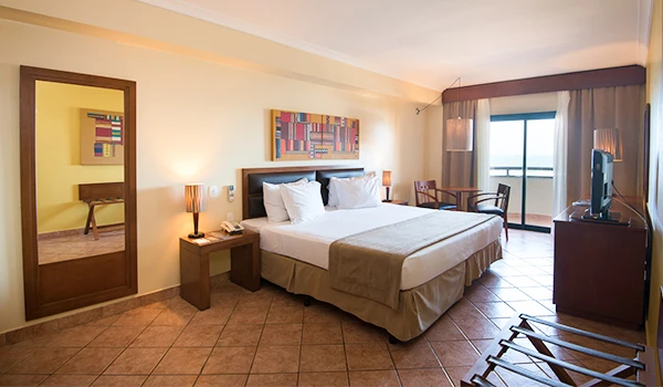 Imagem de um quarto de hotel bem iluminado com cama de casal, duas mesas de cabeceira, televisão e varanda com vista. Hospedagem no Vila Galé Fortaleza.