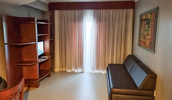 Imagem de um cômodo de hotel com cortinas de tecido, sofá preto de três lugares, estante de madeira com televisão e uma parede decorada, criando um ambiente acolhedor e confortável.
