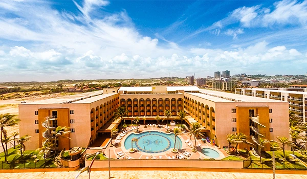 Hospedagem no Vila Galé Fortaleza, foto aérea em que aparece a piscina central ao ar livre, ao redor de jardins bem cuidados, com edifícios de vários andares e um céu azul com nuvens.
