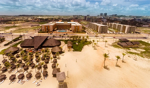 Imagem aérea mostrando resort de luxo na praia com espreguiçadeiras, palhoças de madeira, chão de areia e horizonte com edifícios e céu com nuvens.