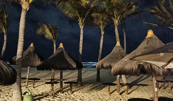 Imagem de uma praia à noite com palapas de sapê, coqueiros ao fundo e céu escuro, transmitindo uma atmosfera tranquila e paradisíaca.