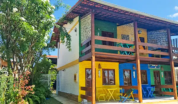 Casa colorida de dois andares com varanda em tons vibrantes de amarelo, verde e azul, cercada por jardim bem cuidado e céu azul claro ao fundo.