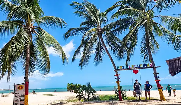 Praia paradisíaca com palmeiras, areia branca, mar azul claro e um arco de entrada decorado em frente ao mar, convidando para relaxar e aproveitar o verão.