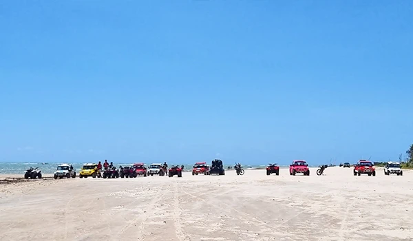 Vários veículos e motocicletas estacionados na praia sob céu azul, com pessoas ao fundo aproveitando o dia de lazer na beira-mar.