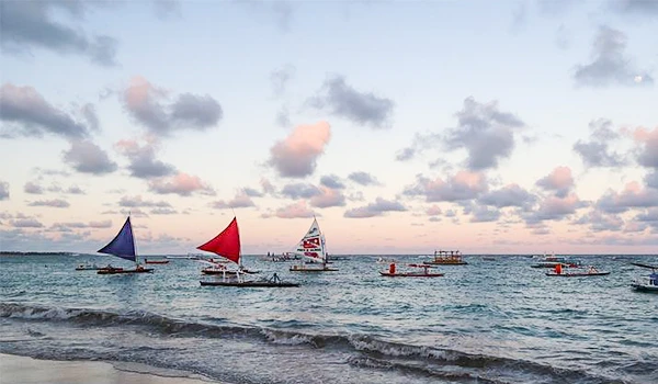 Imagem de um mar na orla com barcos nas cores azul, vermelho e branco, sob um céu com nuvens e uma formação de nuvens de cor rosa ao entardecer