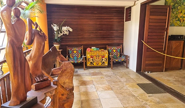 Imagem do interior de uma área de recepção ou lobby decorada com esculturas de madeira, flores e móveis coloridos ao fundo, criando um ambiente acolhedor e bem decorado.