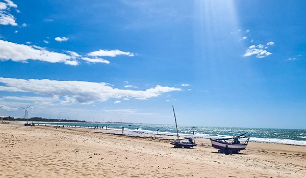 Praia com areia dourada, barcos à vela na orla e céu azul claro com algumas nuvens, ideal para lazer e férias na beira-mar