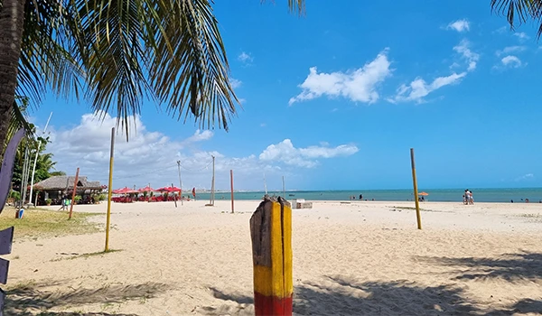 Praia de areia branca com coqueiros, céu azul, mar azul e cabanas na orla, perfeita para lazer e descanso. Observa-se algumas pessoas na praia.