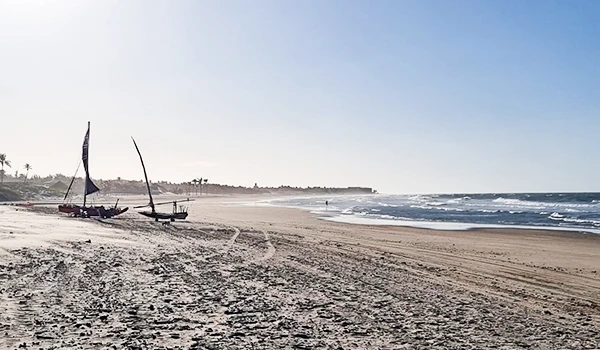 Praia deserta com barcos tradicionais na orla, areia extensa e mar calmo sob um céu claro, perfeito para relaxar e apreciar a natureza.