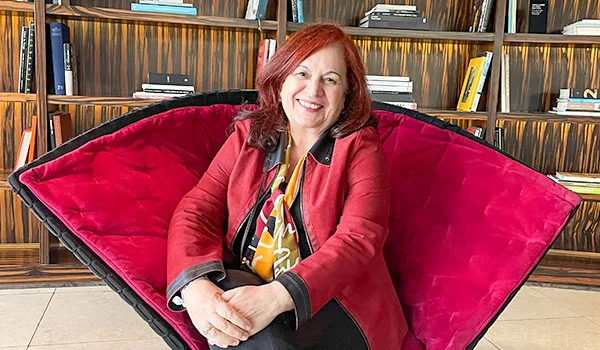 Foto de Deusa Rodrigues em uma poltrona vermelha, exótica com grande espaldar, no lobby do hotel Unique São Paulo, ao fundo uma prateleira com livros. Hotéis de luxo em São Paulo.