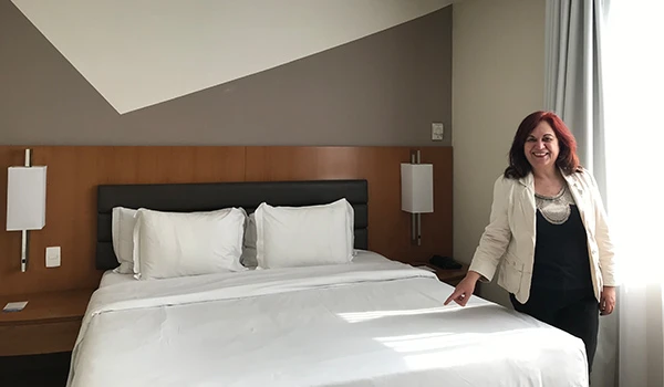 Foto de Deusa Rodrigues no quarto do Hotel Meliá Paulista, onde aparece cama e detalhes da luminosidade da janela.